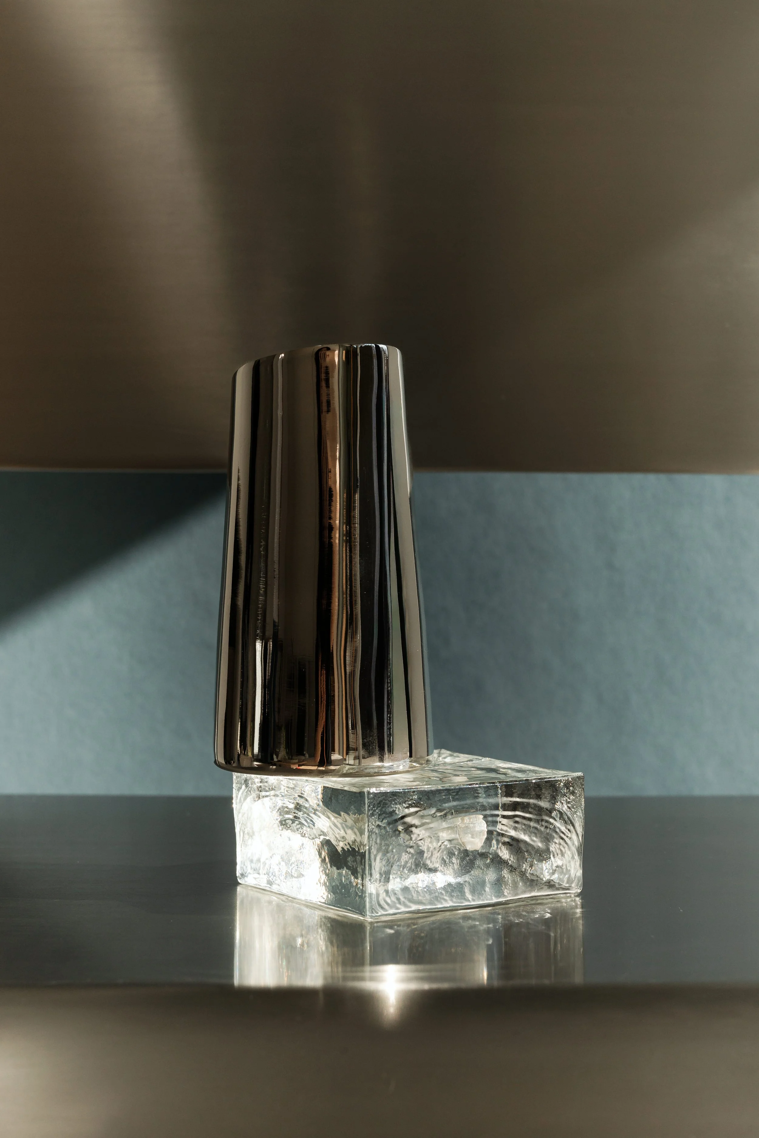 A shiny metallic object placed on a clear glass block, casting reflections and shadows on the surface below. The background is a mix of dark and light shades.