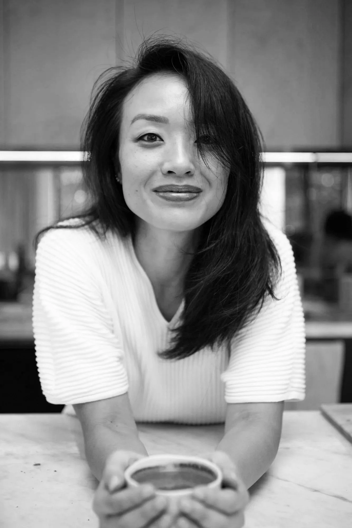 A woman with tousled hair smiling and holding a coffee mug, sitting at a table in a cafe or restaurant, in black and white.