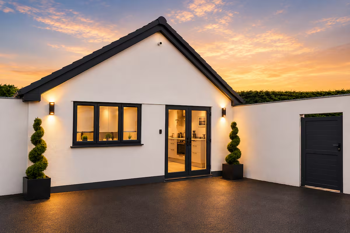 Modern outside view of a small white house with black trim, two potted spiraled bushes, and an open door revealing a bright kitchen, at sunset.