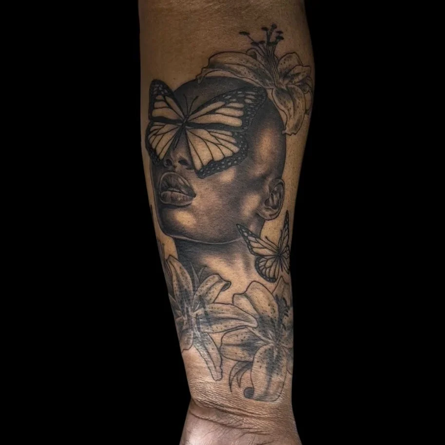 Black and gray tattoo of a woman's face with closed eyes, surrounded by realistic lilies and butterflies on a person's arm.