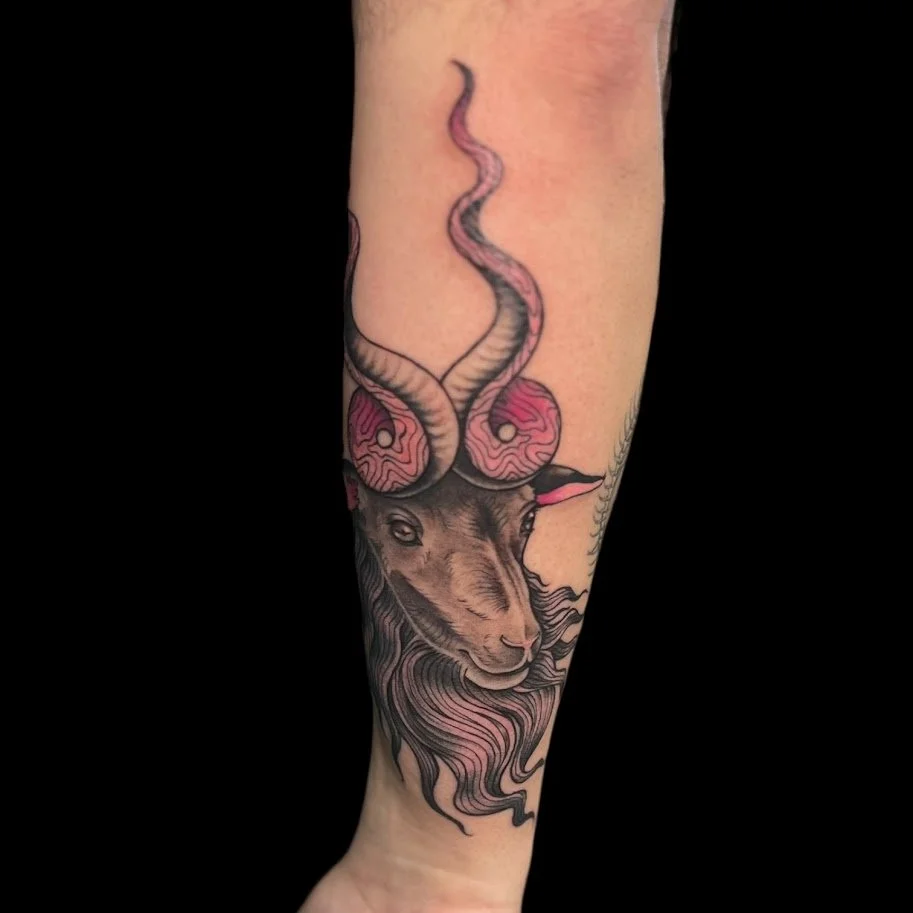 Tattoo of a lion's head with an antelope horned headpiece entwined with pink patterns and black flowing hair on a person's arm.