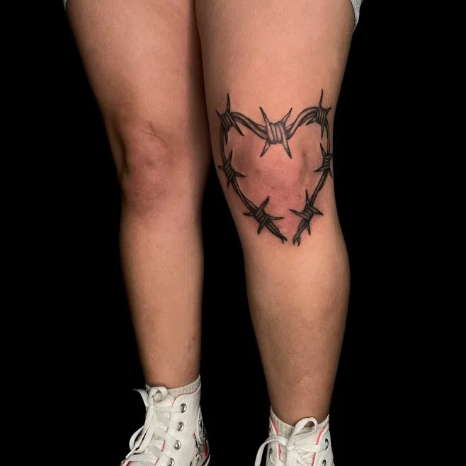 Tattoo of a heart made of barbed wire on person’s right thigh, with the person wearing white sneakers.