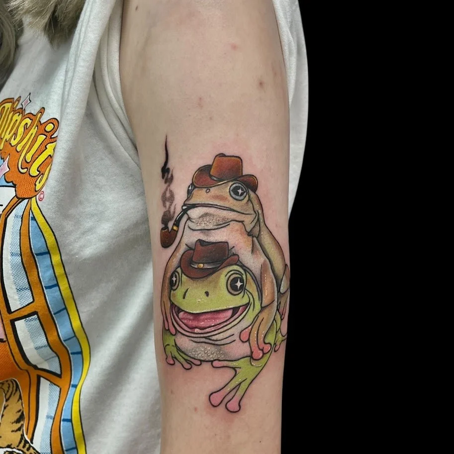 A colorful tattoo of two frogs, one sitting on the other's back, both wearing cowboy hats. The frog on top is smoking a pipe, and the frog below has a wide smile. The design is cartoonish with bold outlines and shading.