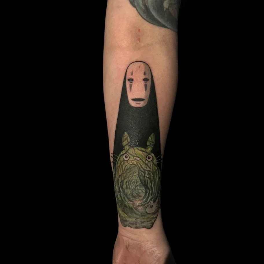 Tattoo of a character with a white face and black tears, resembling No-Face from Spirited Away, combined with a green, textured depiction of a cat at the bottom.