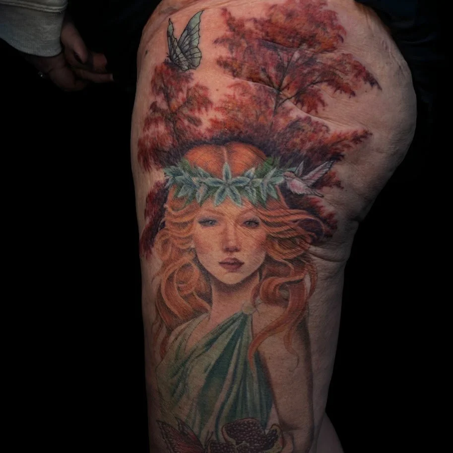 Tattoo of a woman with long red hair, wearing a green dress, surrounded by autumn leaves, with a butterfly and a bird nearby.