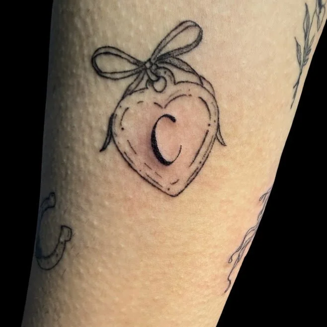 Tattoo of a heart-shaped peach with a letter 'C' inside and a bow on top.