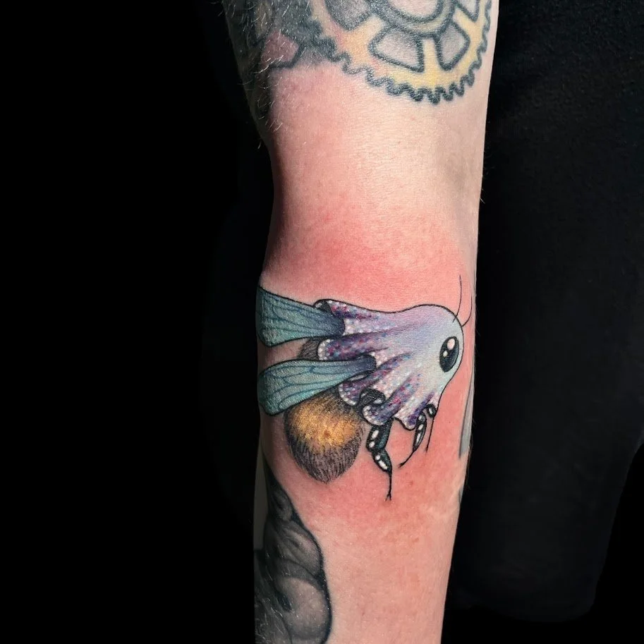 Tattoo of a cartoonish moth with large black eyes and patterned wings on a person's forearm.