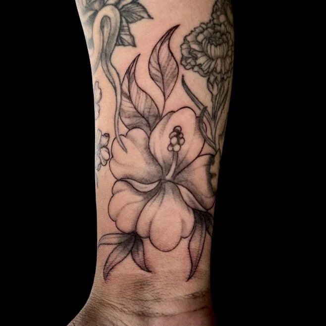 Black and gray tattoo of a large hibiscus flower with leaves on a person's forearm.
