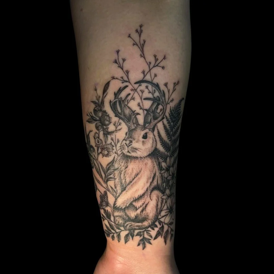 Black and gray tattoo of a cute rabbit with antlers, surrounded by flowers and foliage.