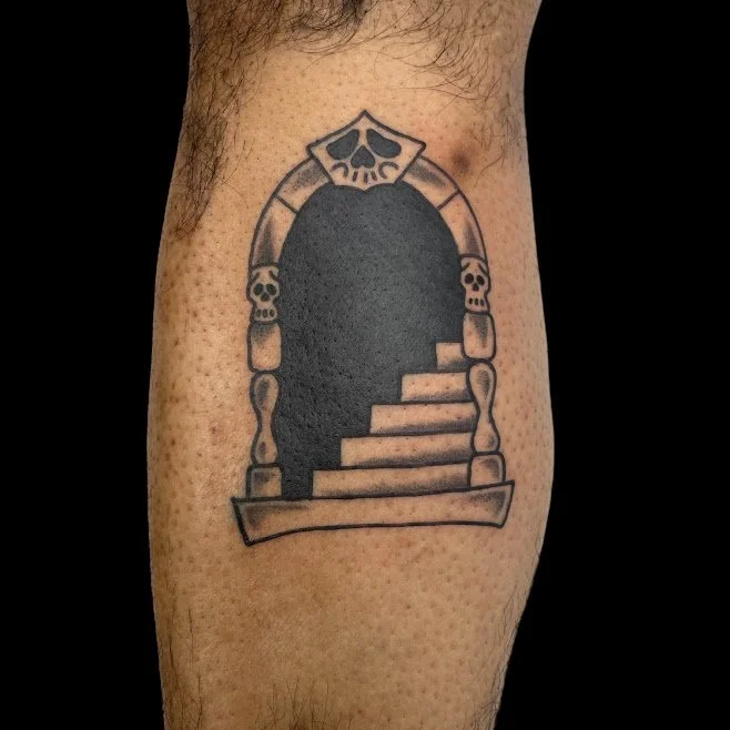 Tattoo of a dark, arched doorway with skull decorations on the pillars and stairs leading into it.