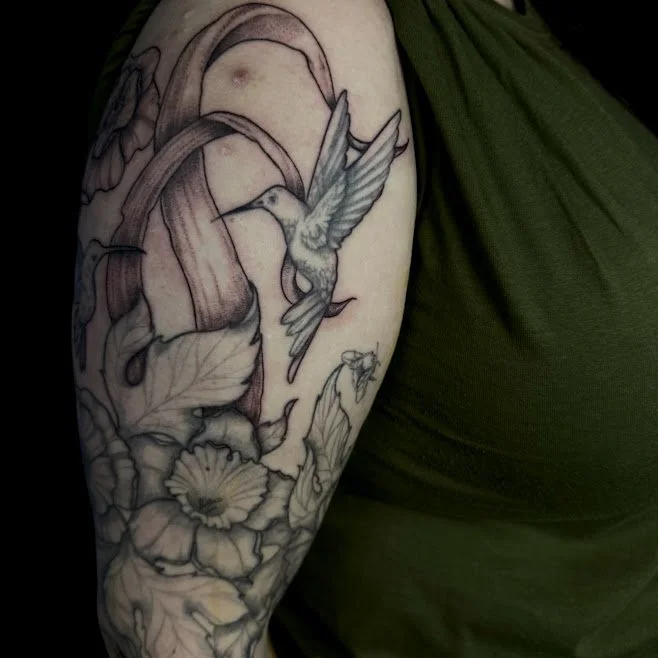 Black and gray tattoo of a hummingbird flying near large flowers and leaves on a person's arm.