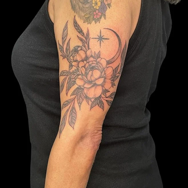 Tattoo of a large peony flower with leaves on a person's upper arm, featuring a crescent moon and star above the flower.