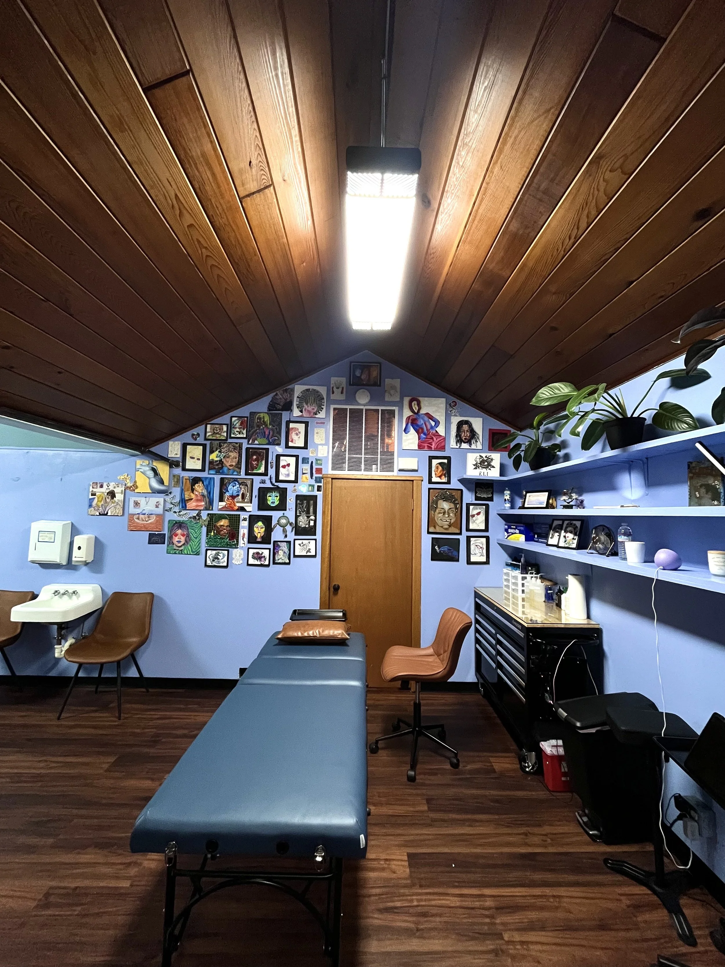 An interior view of a room with a blue wall decorated with numerous colorful artworks and portraits. The ceiling is made of wood panels with a long fluorescent light fixture. There are several chairs, a sink, computer equipment, and potted plants on a shelf, indicating a room used for art or therapy sessions.