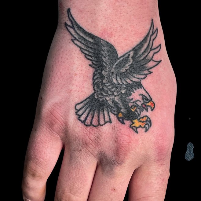 Tattoo of a fierce, detailed hawk with outstretched wings on the top of a person's hand.
