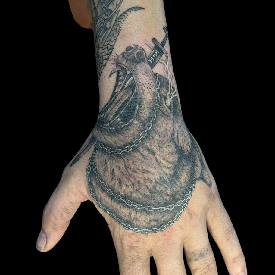 A realistic black and gray tattoo of a roaring bear with its mouth open, covered in chains, on the hand and wrist.