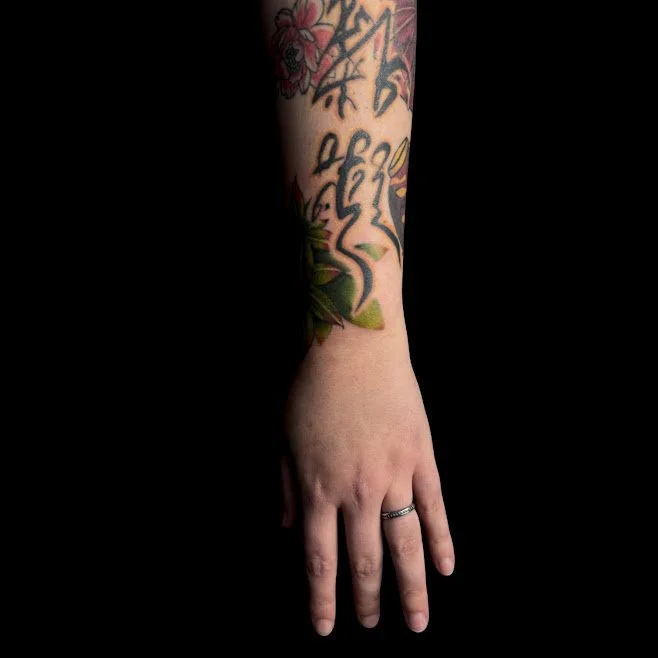 A person's arm with colorful floral tattoos extending from the wrist to the elbow, against a black background, with the hand resting flat and wearing a ring on the ring finger.