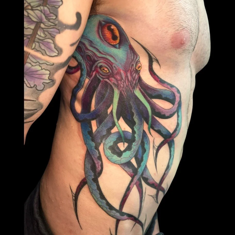 Colorful tattoo of a squid with orange, red, blue, green, and purple hues on a person's side. The squid has large, orange eyes and tentacles extending down the torso.