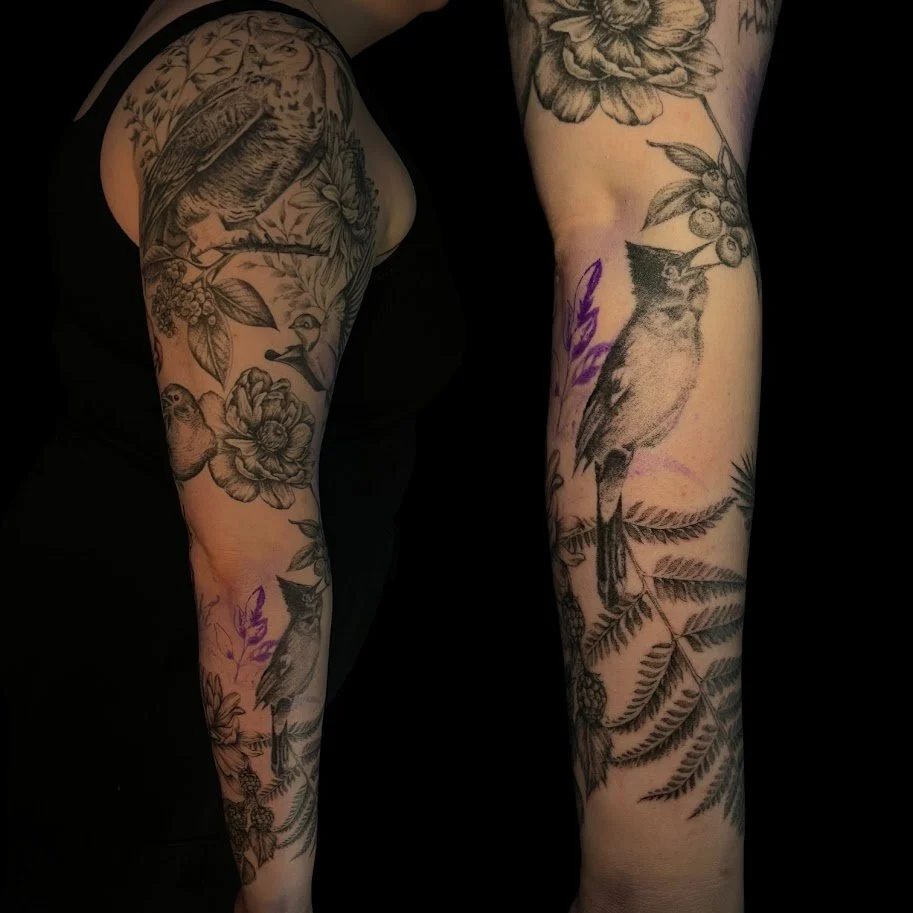 Close-up of a person's arms and shoulder with multiple black and gray tattoos of birds, flowers, and foliage.