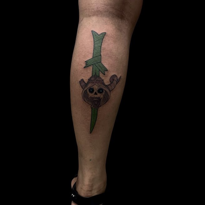 Tattoo of a cartoonish animal face with horns and big eyes on a green spear, with a knife through the spear.