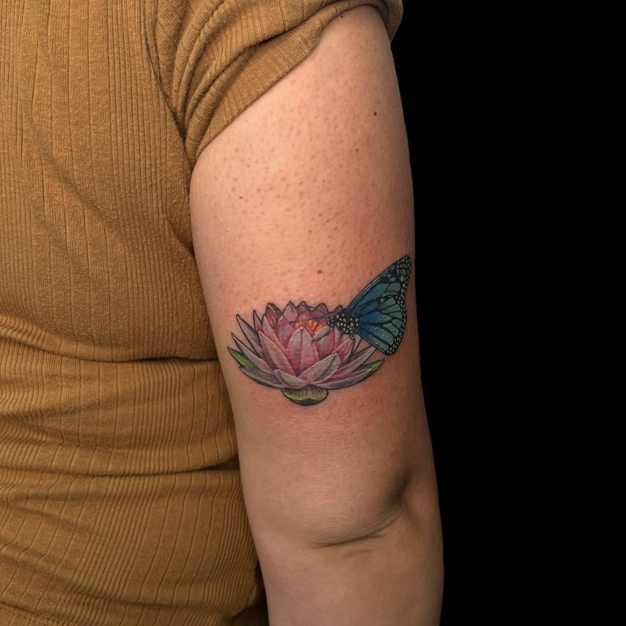 Tattoo of a pink lotus flower with a blue butterfly on its petals on person's upper arm against a black background.