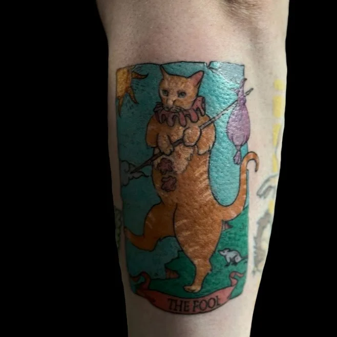 Tattoo of a lion with a cat's head dressed as a fool, holding a stick, with the banner 'The Fool' at the bottom.