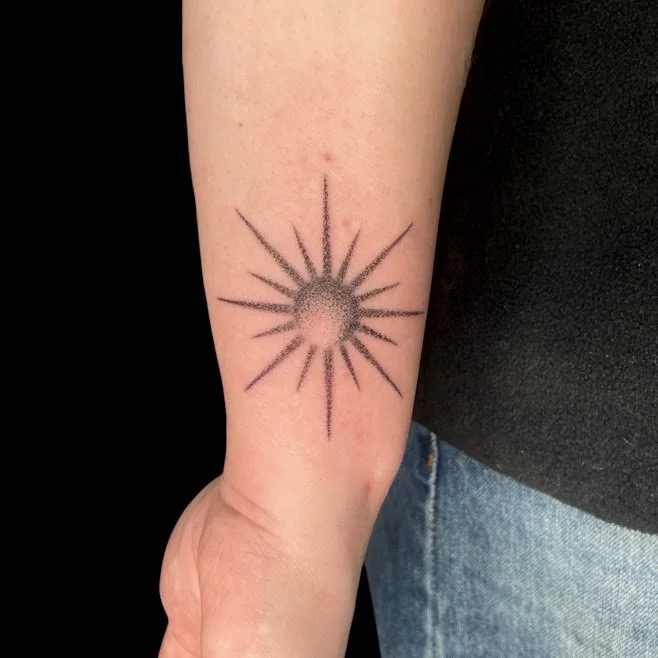 Tattoo of a sun with rays on a person's forearm.