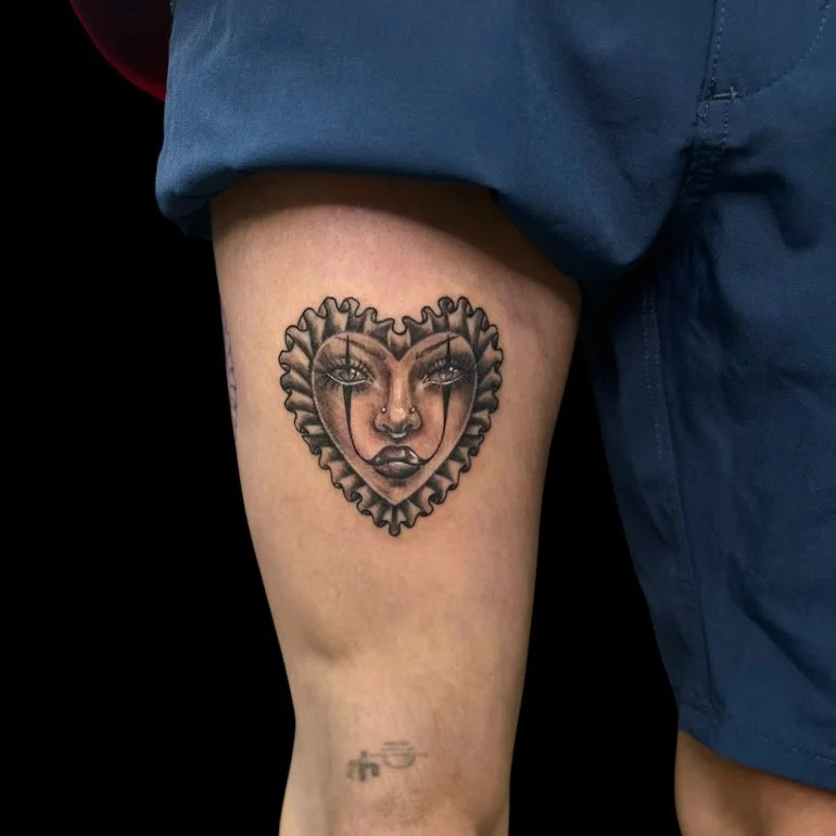 Tattoo of a woman's face framed by a heart-shaped design on a person's thigh.