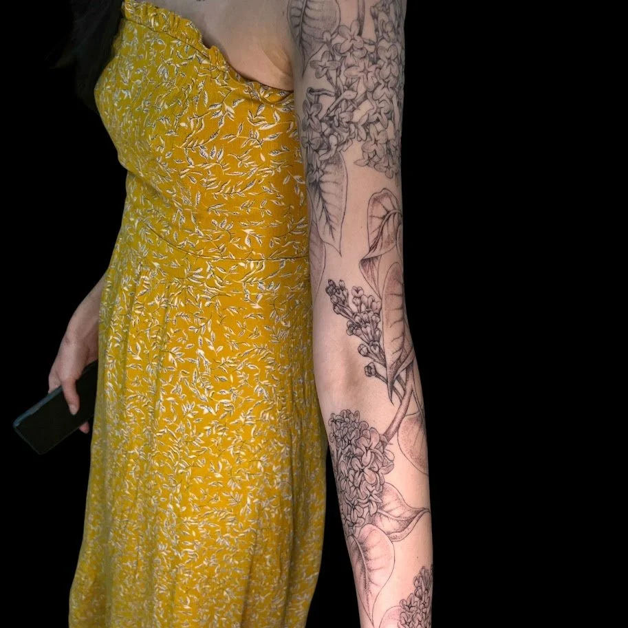 A woman wearing a yellow floral dress holding a phone, with her tattooed left arm exposed, standing against a black background.