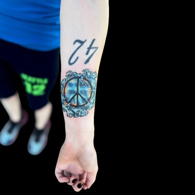 Tattoo of a peace sign surrounded by a decorative blue and gray design on the person's forearm, with the number 42 written above.