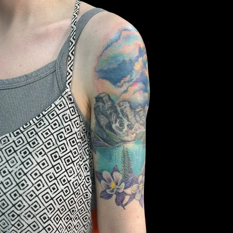 Colorful tattoo sleeve featuring mountains, a lake, a waterfall, and flowers on a person's upper arm.