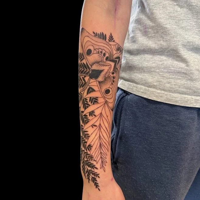 A person's arm with a detailed black and gray tattoo featuring abstract geometric shapes and leaf patterns, extending from the wrist to the elbow.
