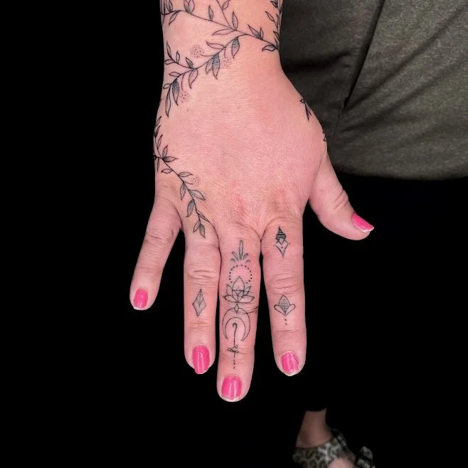 A person with pink nail polish showing their decorated hand and fingers with various small black tattoos and a sleeve tattoo of leafy vines on their arm.