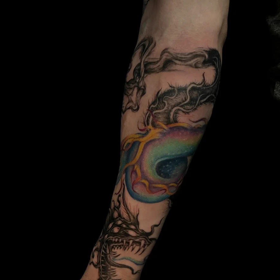 Colorful tattoo of a galaxy or black hole with swirling shapes, smoke, and a dragon on a person's arm.
