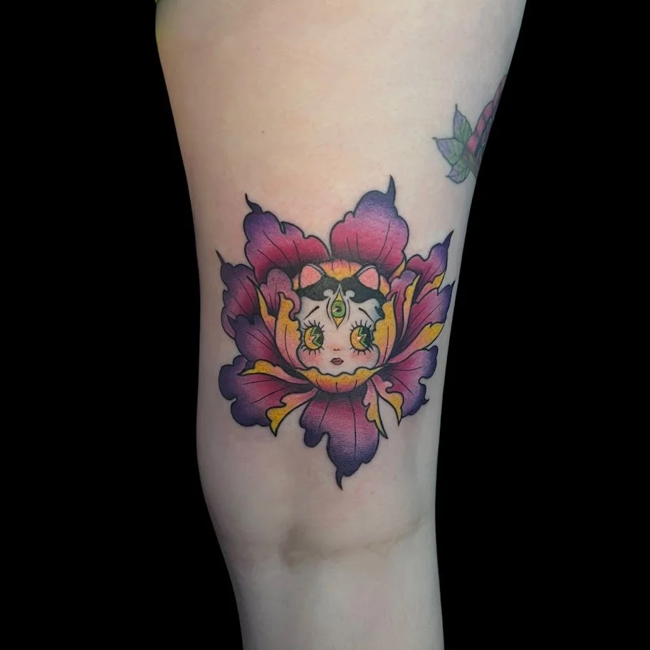 Tattoo of a stylized animal face with big eyes and a third eye on its forehead, surrounded by purple and pink leaf-like shapes on a person's arm.