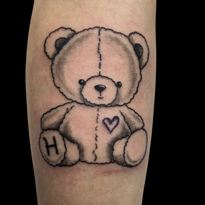 Tattoo of a cute teddy bear with a heart on its chest and the letter H on one foot, sitting on skin.