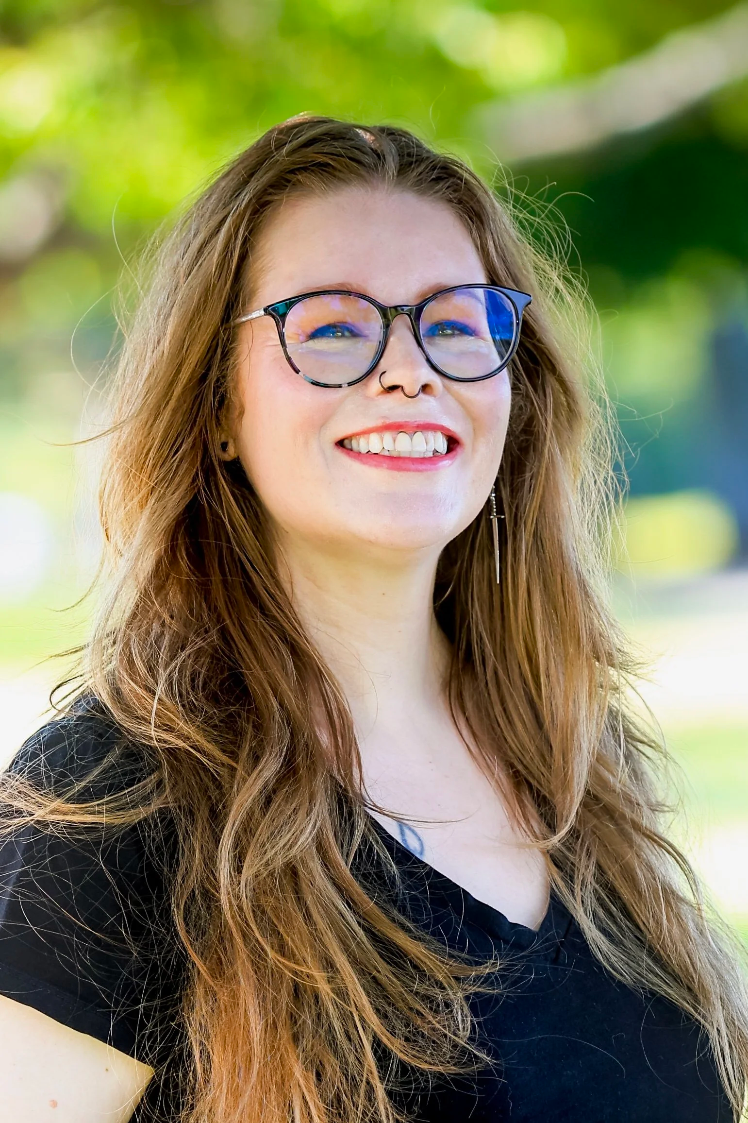 A young woman with long, curly red hair wearing glasses with a colorful frame, smiling outdoors with a blurred green leafy background.