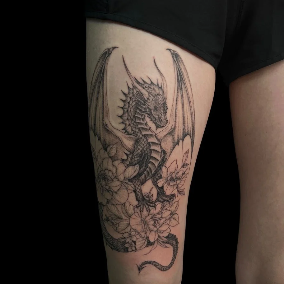 A detailed black and gray tattoo of a dragon on a person's thigh, surrounded by flowers.