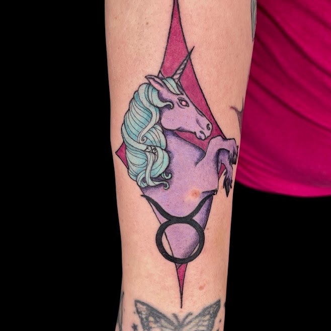 Tattoo of a purple unicorn with a teal mane on a person's arm, with a pink background and a black circle symbol below.