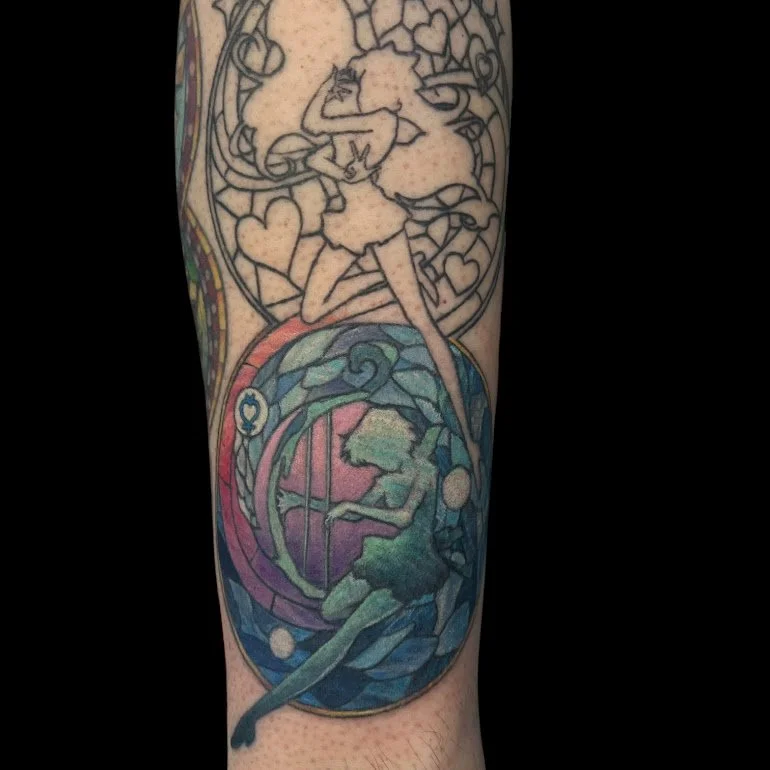 A colorful tattoo featuring two distinct sections. The upper part depicts a cartoon-style character with long hair, a dress, and a playful pose, surrounded by hearts. The lower part showcases a more detailed, fantasy-style scene with a figure, possib