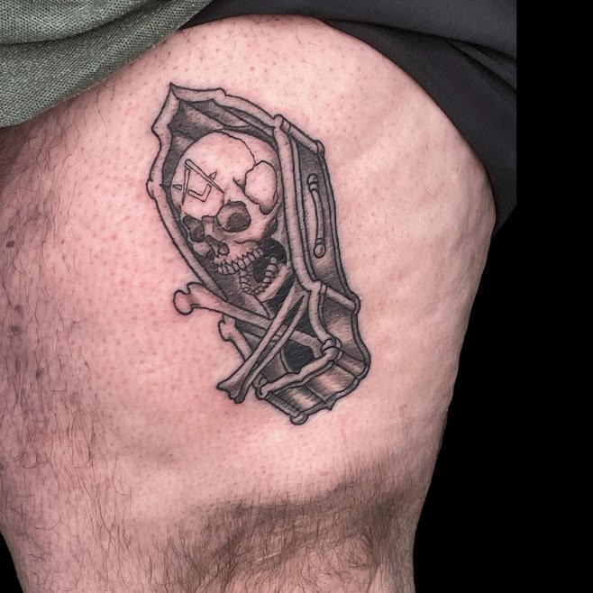 Tattoo of a skull inside a coffin with crossed bones and a wrench on the side, on upper thigh.
