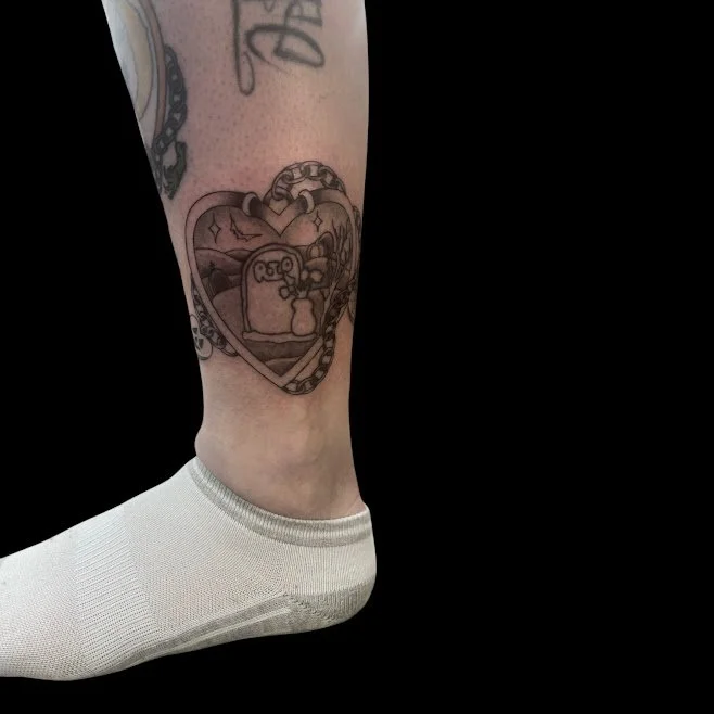 A person's lower leg and foot, with a heart-shaped tattoo featuring a cartoon character holding a cat, surrounded by smaller designs, against a black background.