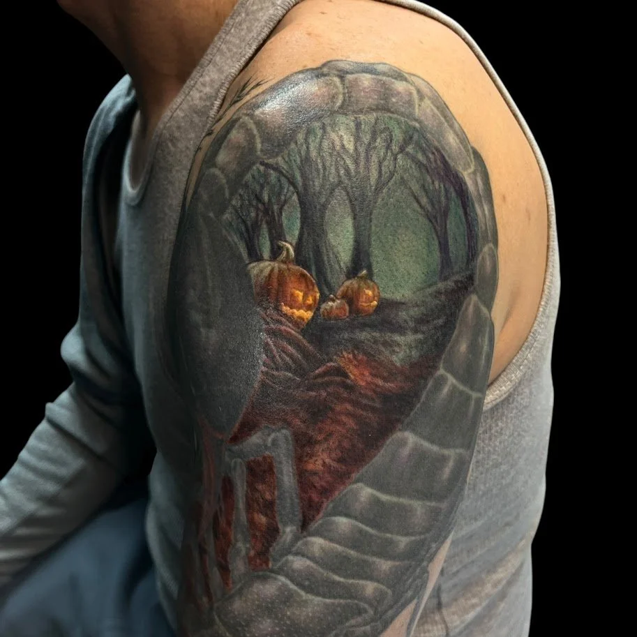 Tattoo of a pumpkin patch with glowing jack-o'-lanterns on a forest path, framed by a stone archway on the person's arm.