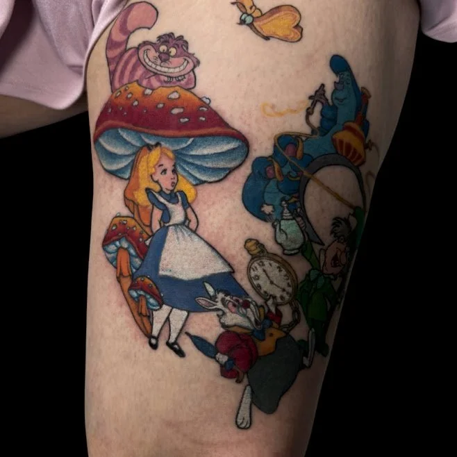 Tattoo of characters from Alice in Wonderland, including Alice, the White Rabbit, the Mad Hatter, and the Cheshire Cat, on a person's thigh.