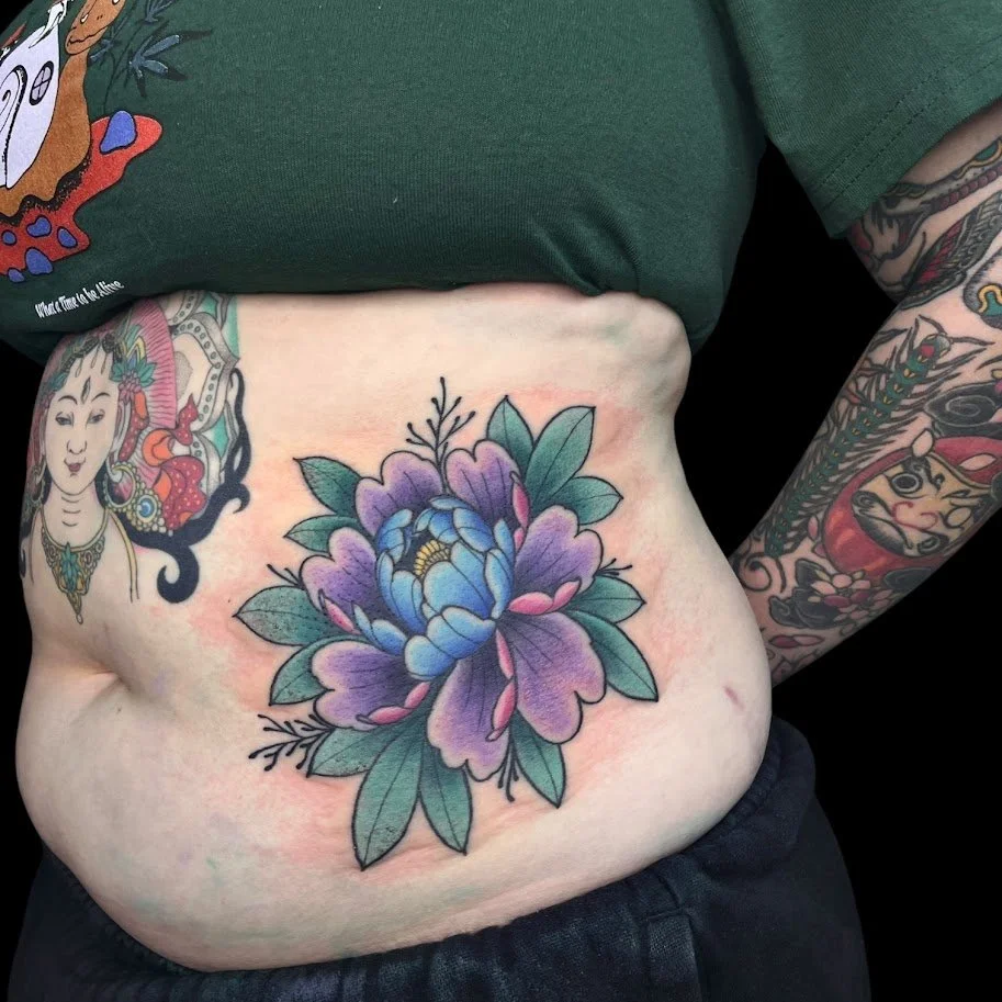 Colorful floral tattoo on person's stomach, featuring purple and blue flowers with green leaves, and tattooed arm with multicolored designs.