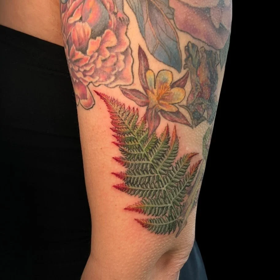Tattoo of a green fern leaf with red tips, surrounded by other colorful floral tattoos on the person's upper arm.