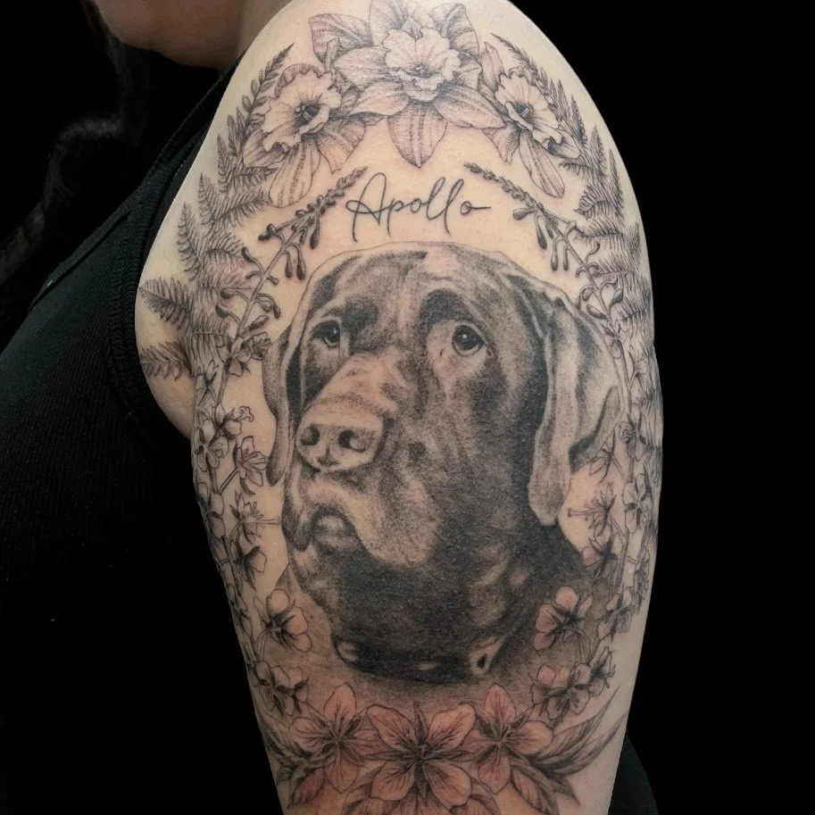 Black and gray tattoo of a dog's head surrounded by flowers and leaves, with the word "Apollo" written above.