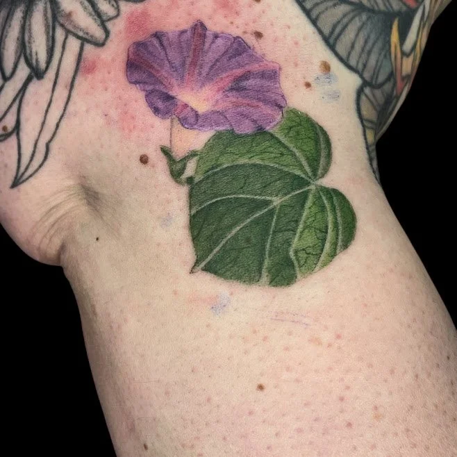 Close-up of a tattoo of a green ivy leaf and a purple morning glory flower on a person's arm.