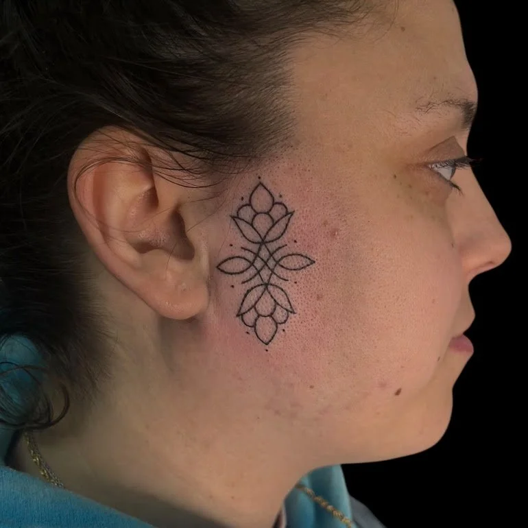 Side profile of a woman with a geometric floral tattoo on her cheek near her ear, wearing a blue top and a gold necklace, against a black background.