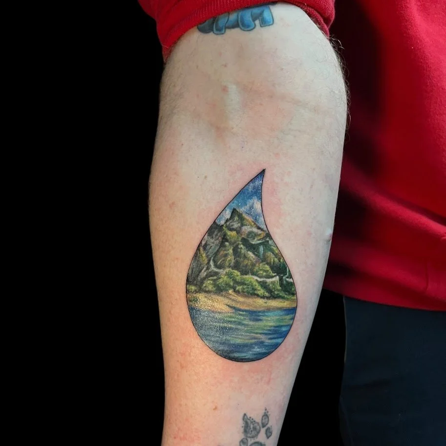 Tattoo of a landscape within a teardrop shape on a person's upper arm, featuring mountains, trees, and a river.