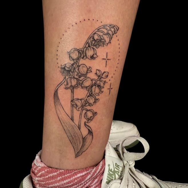 Black and gray tattoo of lily of the valley flowers on a person's leg, with dotted circle and cross marks around it.